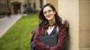 A smiling young woman holding a laptop leaning against an outdoor university building