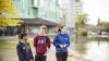 Three students (two young men and a young woman) walking outdoors by a river on the grounds of their university. They are dressed casually in hoodies and appear to be engaged in conversation. University buildings are visible in the background.