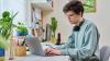 Young man wearing headphones using a laptop at a desk surrounded by plants and stationery